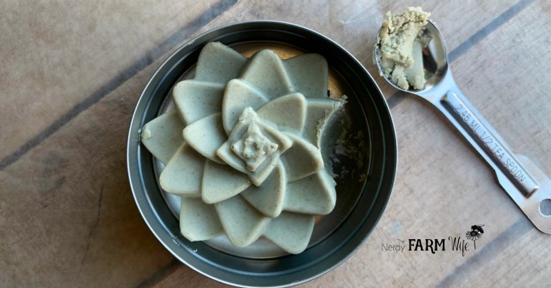 green clay cleansing bar in a metal tin, with a small half teaspoon beside it