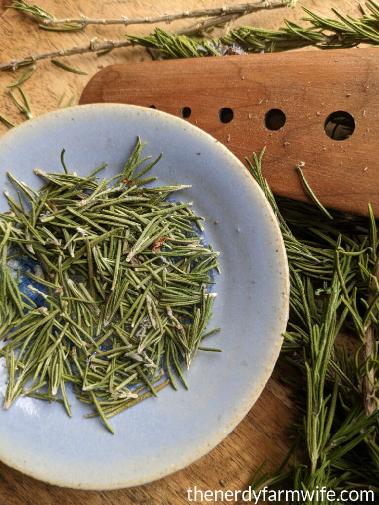 plate of rosemary leaves next to a wooden herb stripper.